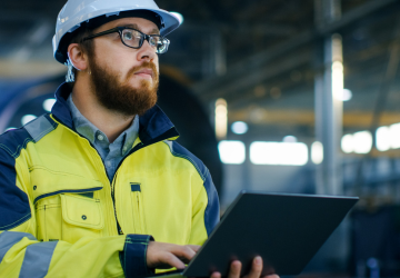 Industrial Engineer in Hard Hat Wearing Safety Jacket Uses Laptop. He Works in the Heavy Industry Manufacturing Factory with Various Metalworking Processes are in Progress.