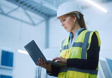 Professional Female Engineer Wearing Safety Jacket and Hard Hat Holding and Working on Laptop Computer. Modern Bright Industrial Facility.