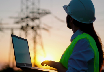 Woman engineer, power engineer in helmet checks power line using computer laptop online. Electric lines of high voltage at sunset.